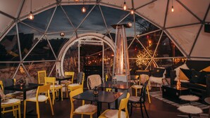 Una estructura en forma de cúpula geodésica con mesas y sillas dispuestas en su interior, que cuenta con un elemento calefactor central e iluminación ambiental.
