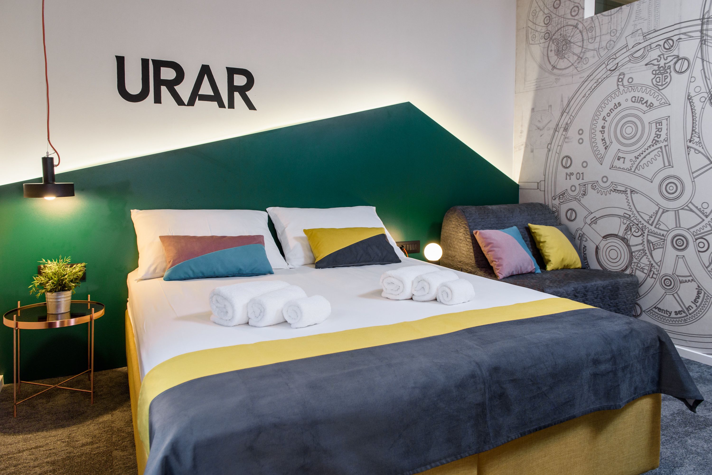 A neatly made bed with colorful pillows and a green headboard. A small table with a plant and a lamp is visible.