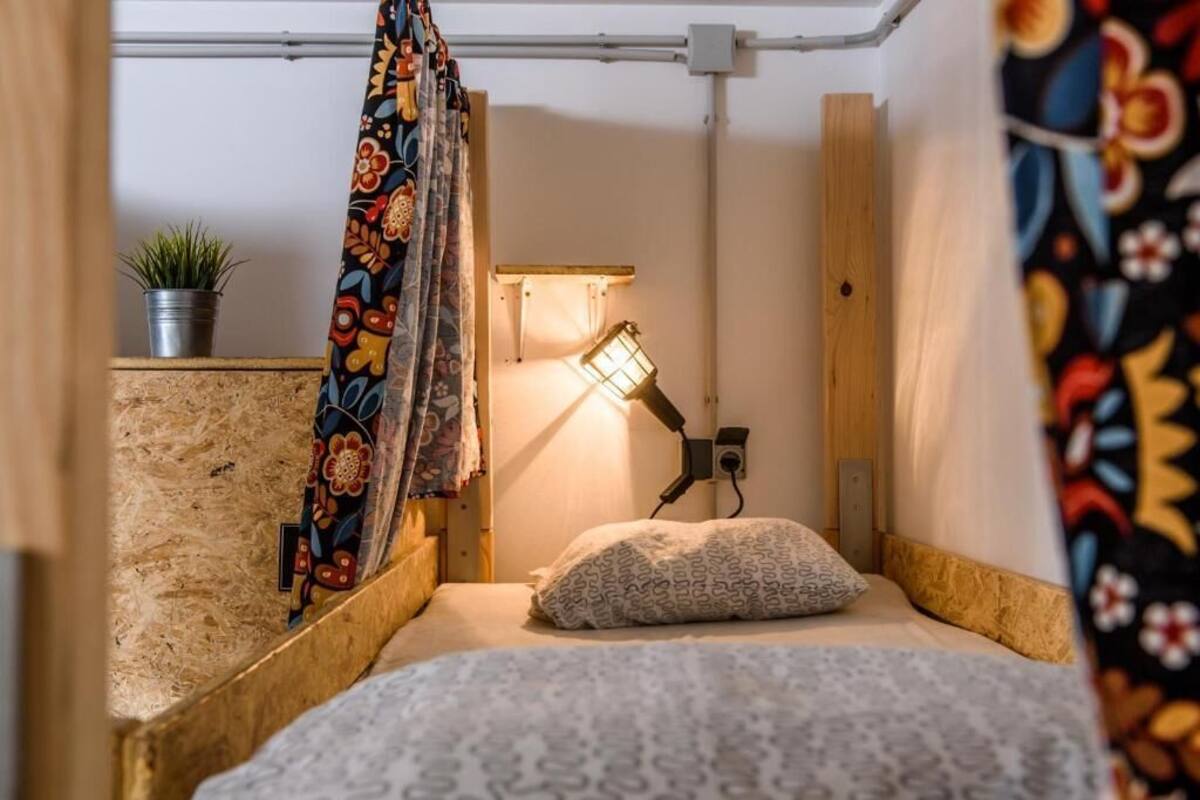 Ein kleiner, gemütlicher Raum mit einem Bett, einer Wandlampe, einem Topf mit Pflanze und einem farbenfrohen Vorhang.