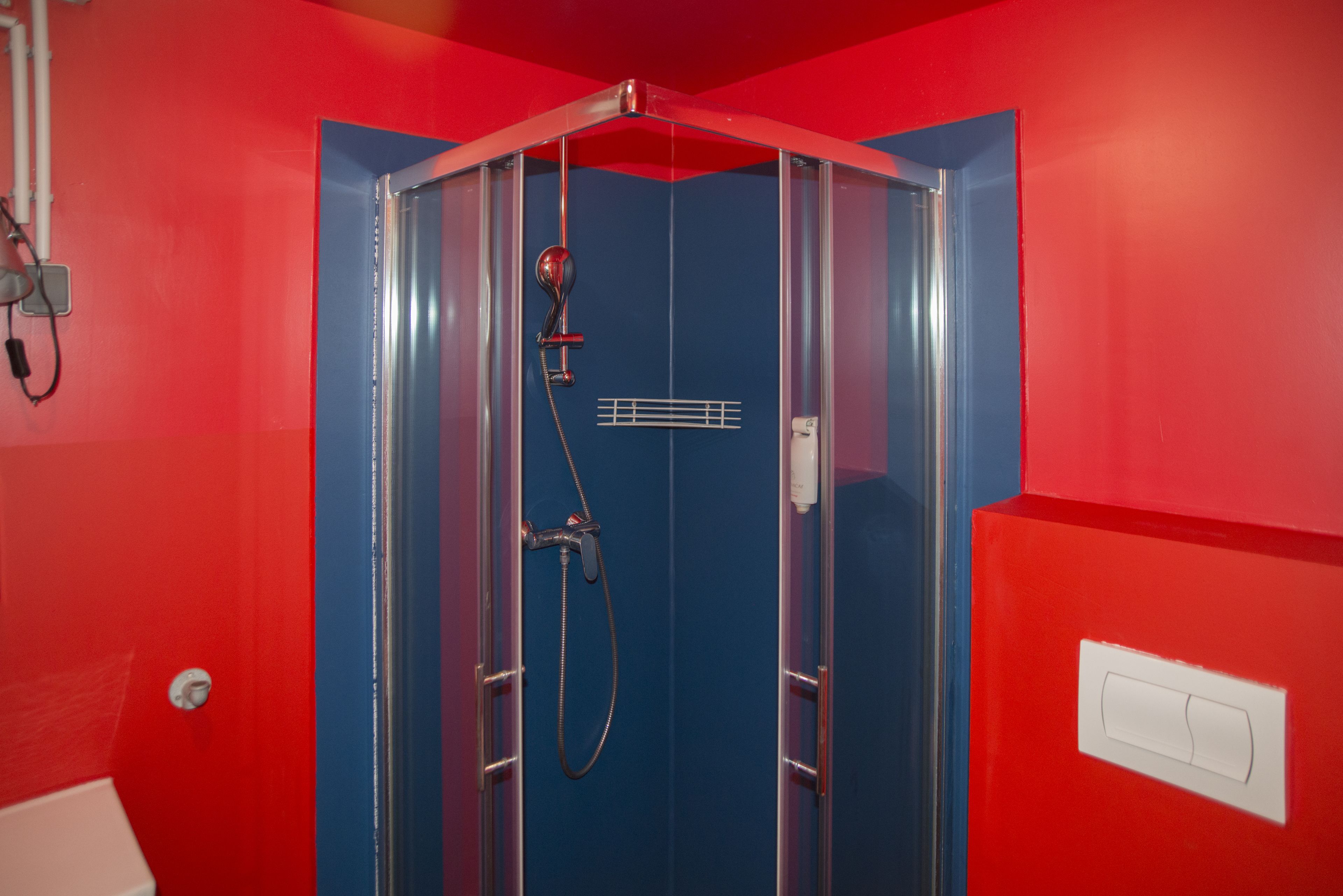 A room with a red wall, a shower with a glass door, and a white switchboard.