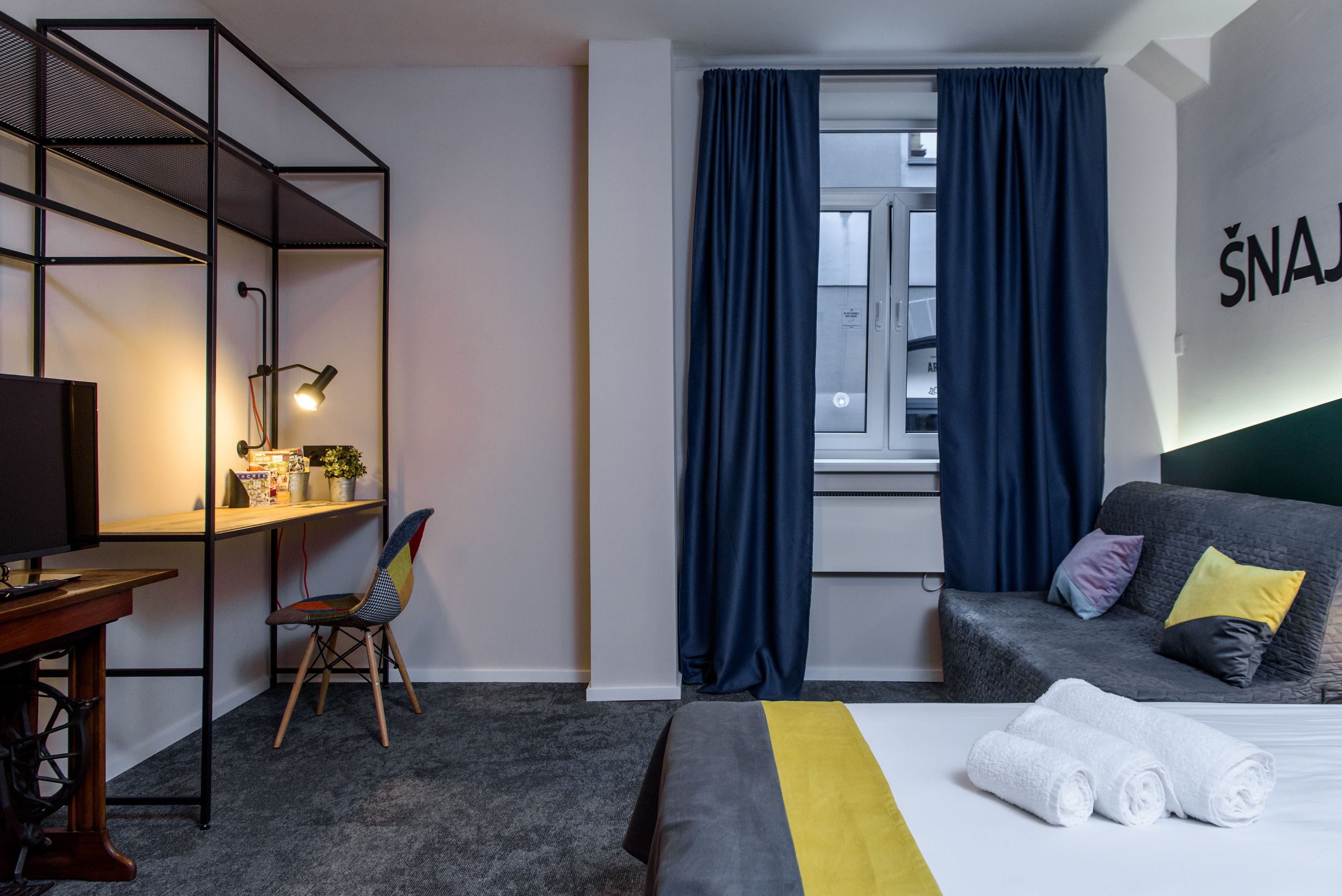 Habitación de hotel con cama, escritorio, silla, estantería y ventana con cortinas.