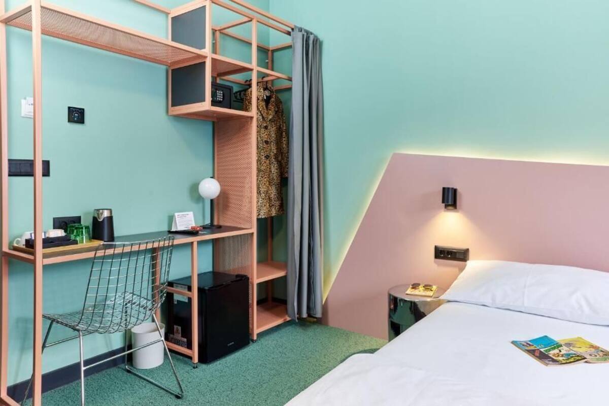 Ein kleines Hotelzimmer mit Bett, Schreibtisch und Stuhl, einem kleinen Tisch, einem Kleiderschrank und einem Nachttisch mit Lampe.