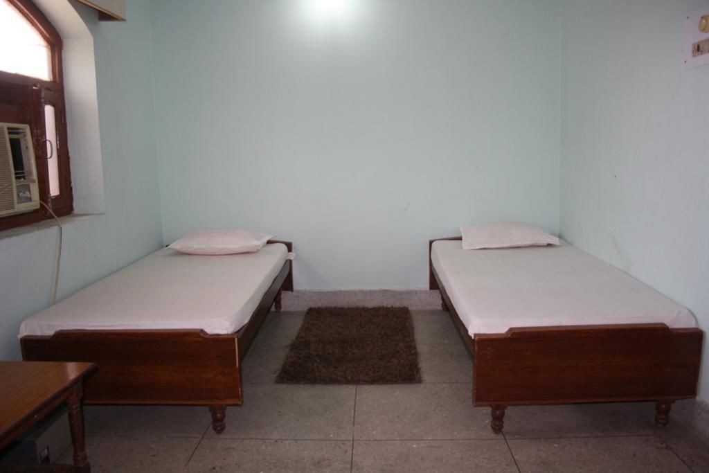Une petite pièce simple, avec deux lits en bois, un petit tapis et une fenêtre équipée d’un appareil de climatisation.