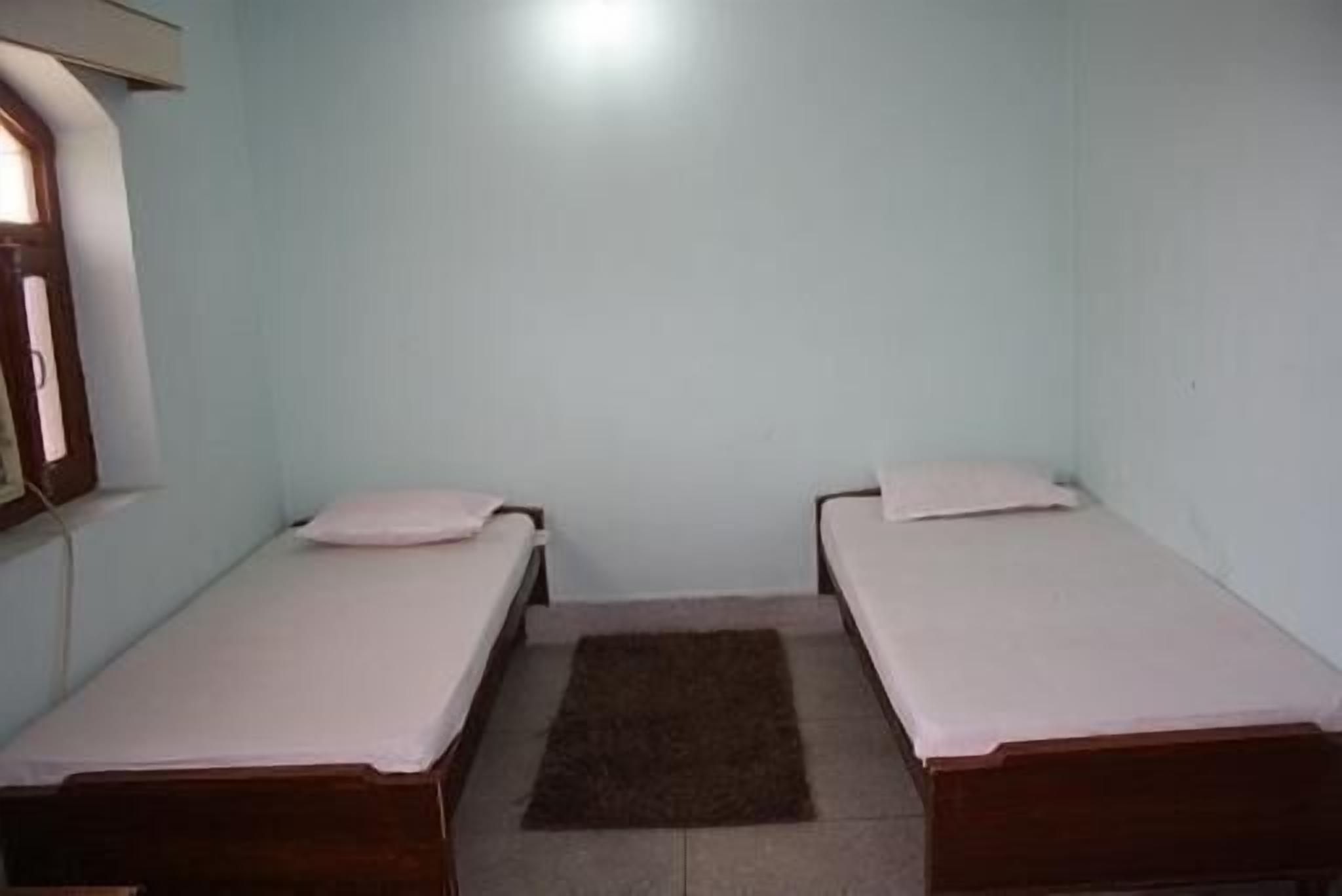 A small, single-room with two single beds, a window on one wall, and a brown rug on the floor.