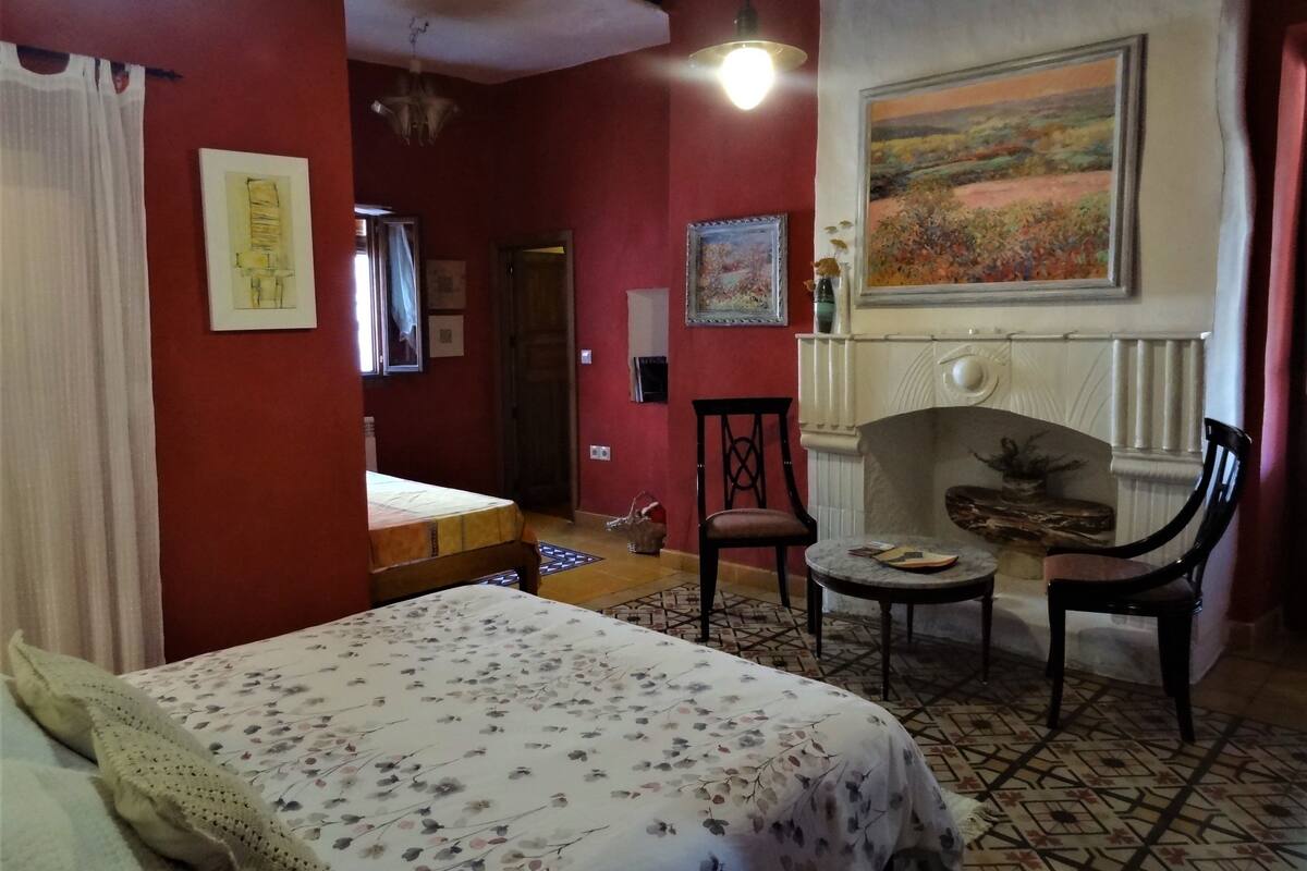 Une pièce comprenant un lit, une chaise, une petite table, une cheminée et un tableau accroché au mur.