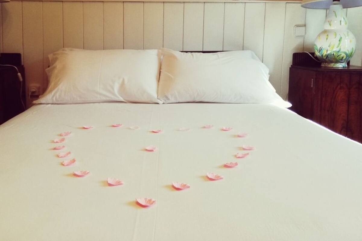 Un lit recouvert de draps blancs, et sur le couvre-lit, une composition en forme de cœur faite de pétales de roses.