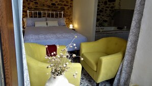 A bedroom with a stone wall, a bed with a blue bedspread, two yellow armchairs, and a vase with flowers. - Hostal La Solana (Arenas de San Pedro)