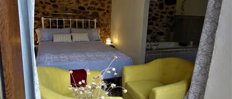 A bedroom with a stone wall, a bed with a blue bedspread, two yellow armchairs, and a vase with flowers.