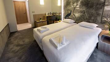 A hotel room with two beds, a desk, a chair, a small table with a plant, and a peacock mural on the wall.