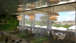 A restaurant with tables set for dining, large windows offering a city view, and chandeliers hanging from the ceiling. - Park Hotel RAYA Garden (Veliko Tarnovo)