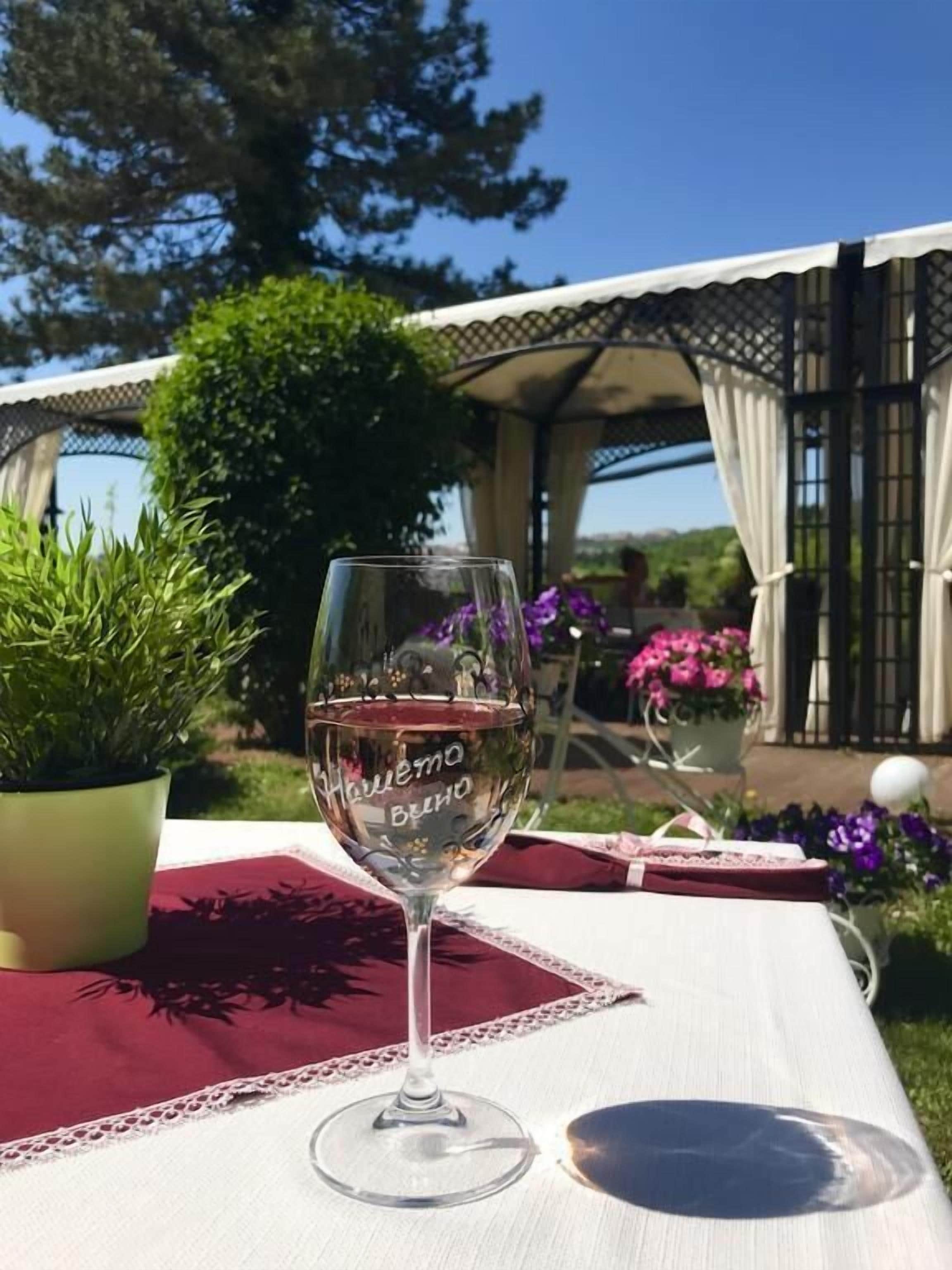 A glass of white wine on a table with a lace table runner, surrounded by greenery and flowers.