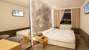 A hotel room with two beds, a desk, a chair, a TV, and a city view through the window. - Park Hotel RAYA Garden (Veliko Tarnovo)