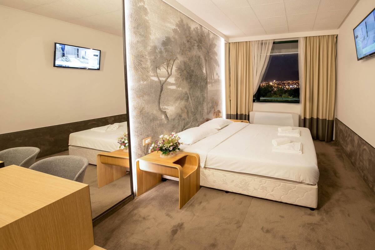 A hotel room with two beds, a desk, a chair, a TV, and a city view through the window.