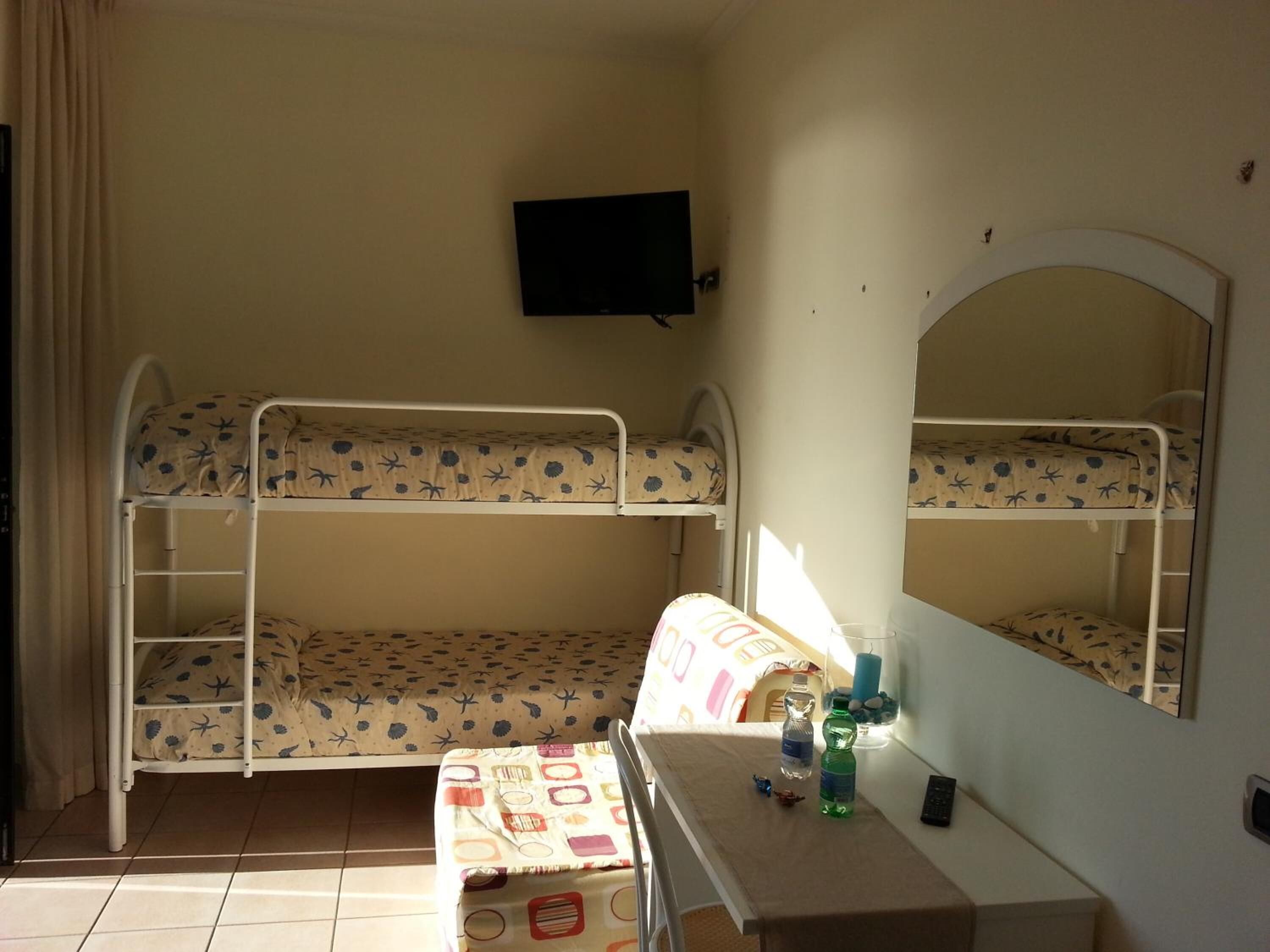 A bunk room with two bunk beds, a desk with a mirror, a television on the wall, and a window with curtains.