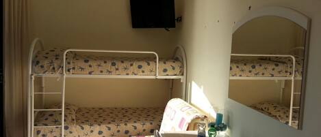 A bunk room with two bunk beds, a desk with a mirror, a television on the wall, and a window with curtains.