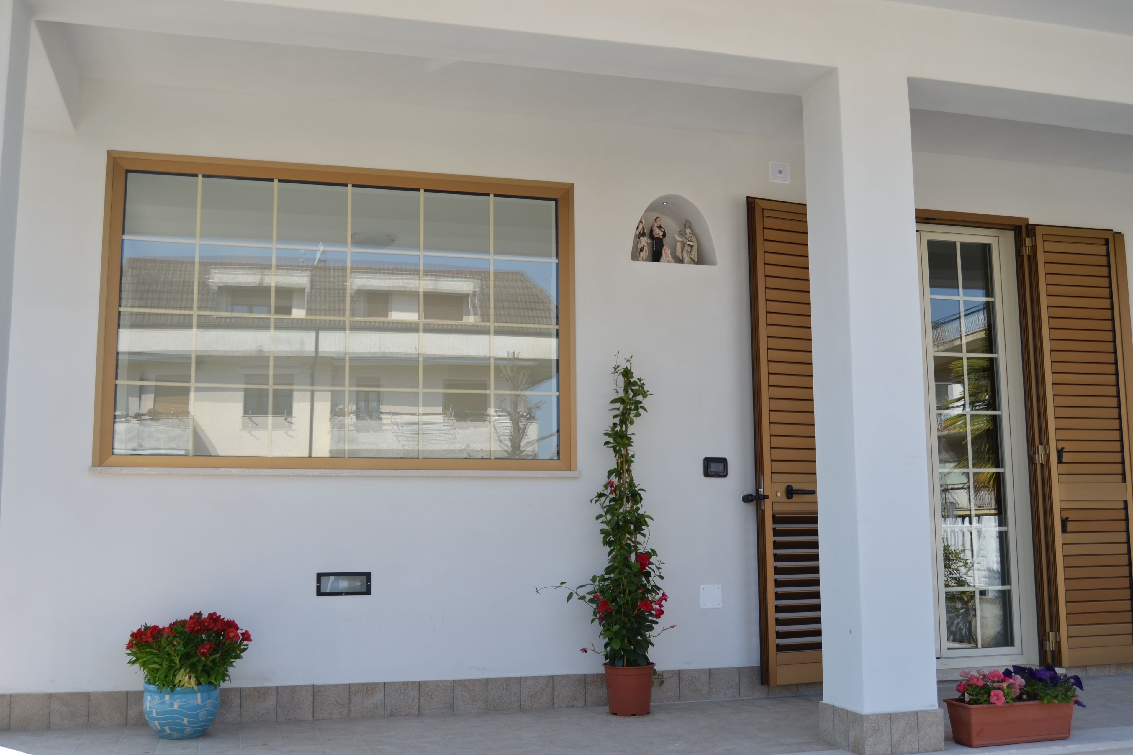 Fachada de edificio con un amplio ventanal, una puerta con contraventanas de madera y dos macetas con flores.