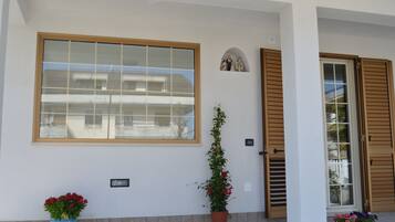 Fachada de edificio con un amplio ventanal, una puerta con contraventanas de madera y dos macetas con flores.