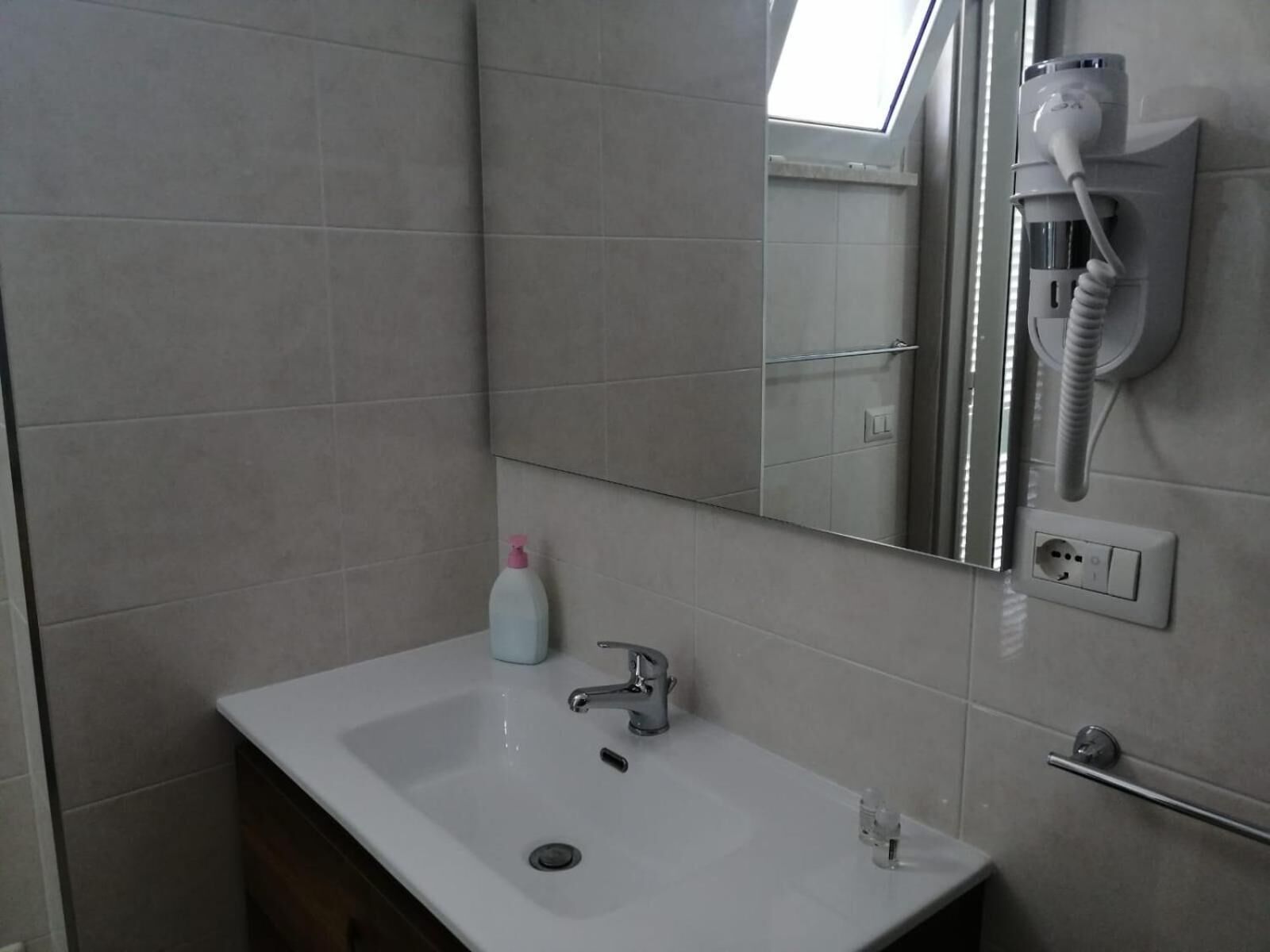 Une salle de bain avec un lavabo blanc, un miroir, un sèche-cheveux fixé au mur et un flacon de savon liquide.