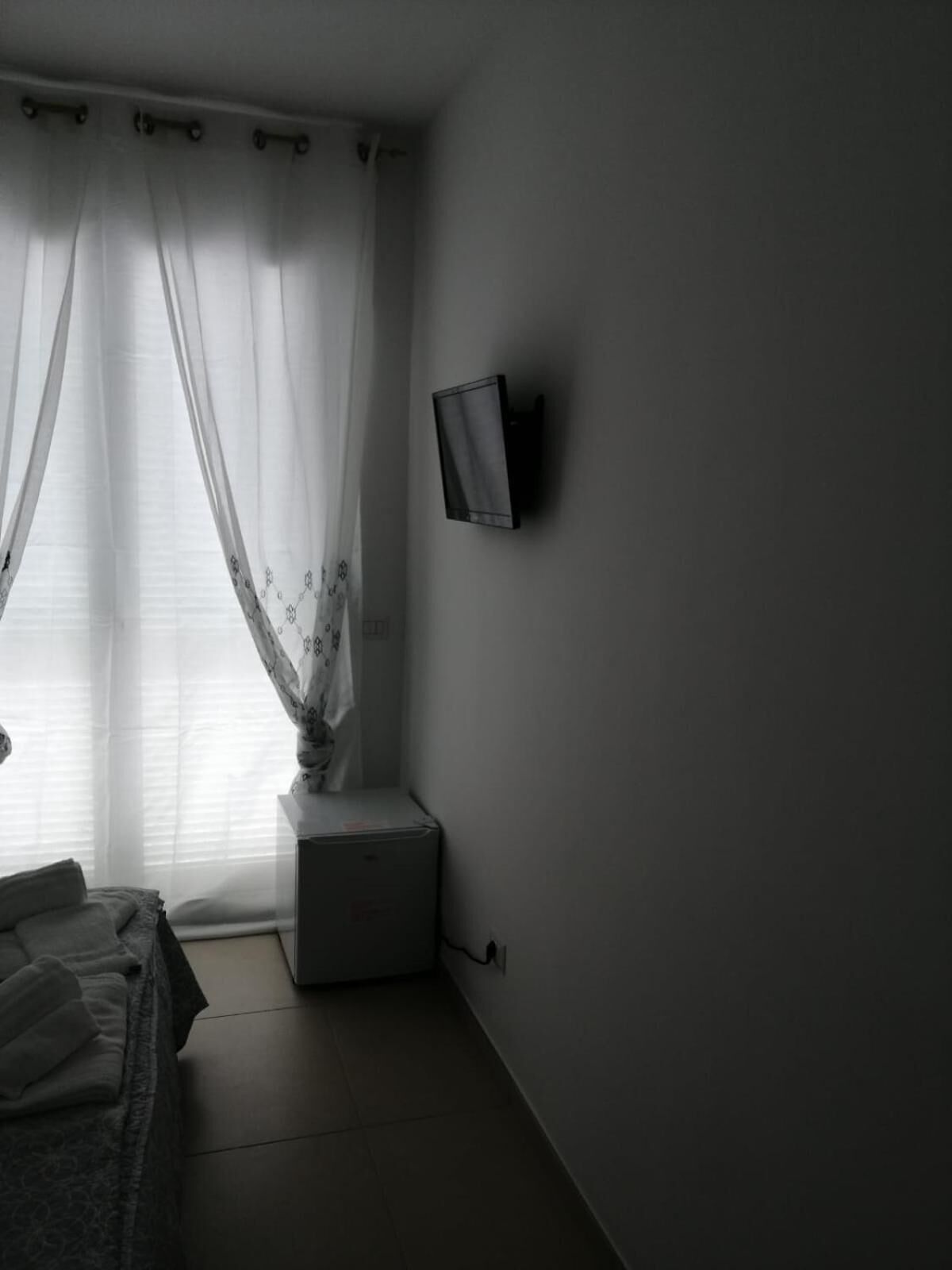 Una habitación tenuemente iluminada con una cama, una mesita de noche pequeña, un televisor montado en la pared y cortinas traslúcidas.