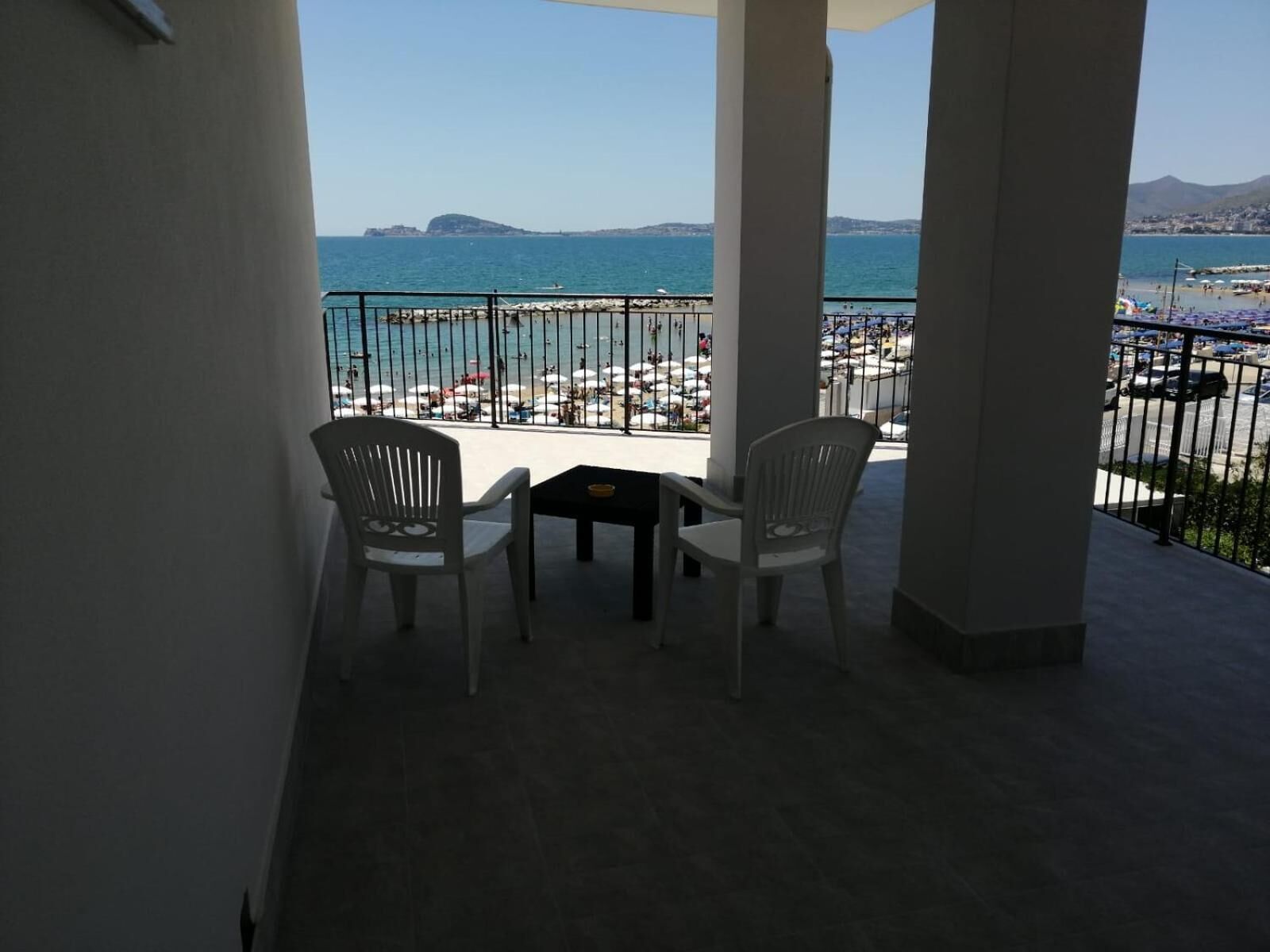A balcony with two chairs and a small table, overlooking a beach with umbrellas and a clear view of the sea and distant mountains.