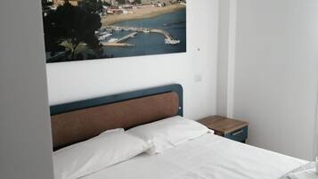 Un dormitorio con cama, mesita de noche, cuadro de un pueblo costero en la pared y una ventana.