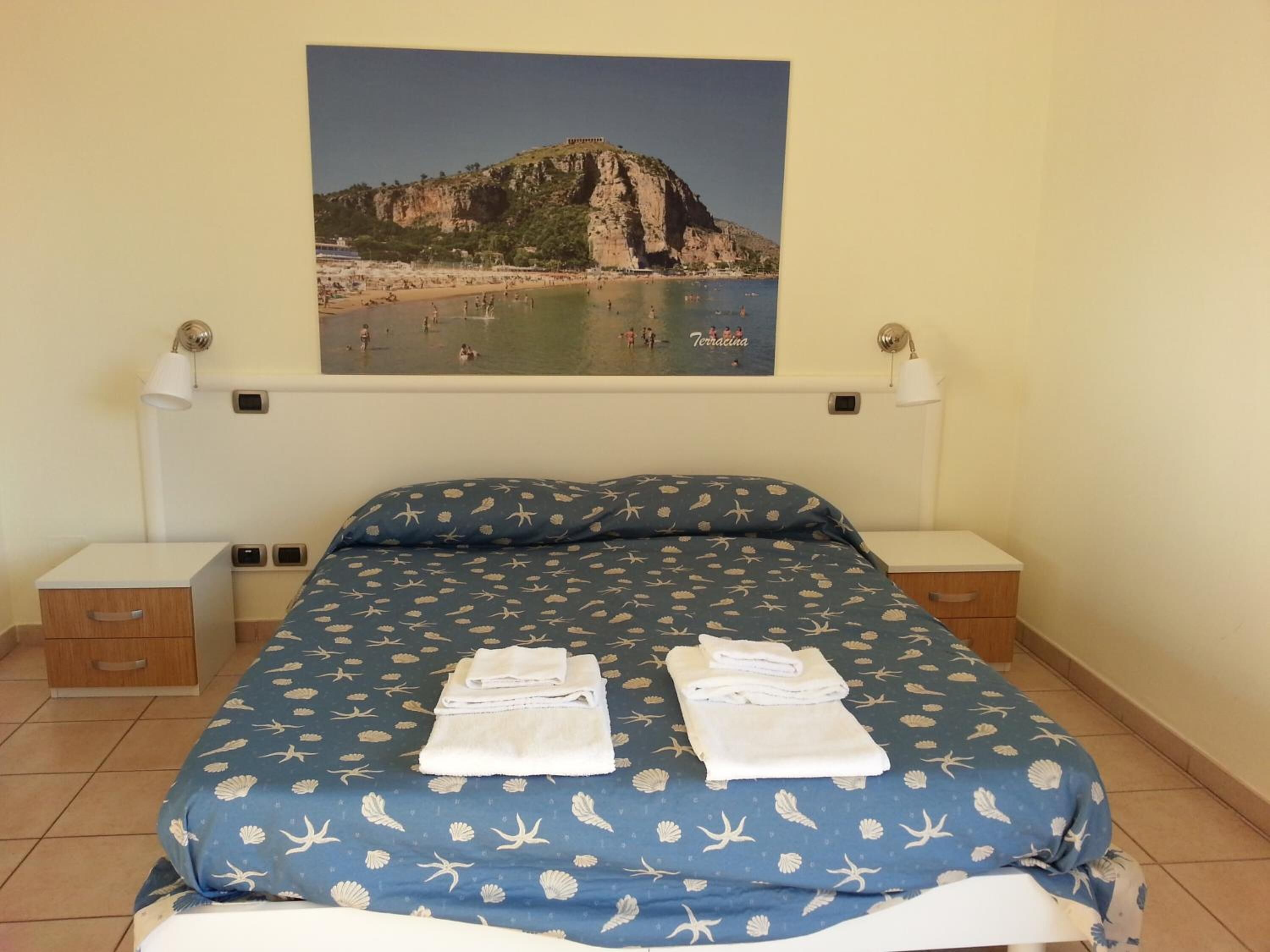 Un dormitorio con una cama, dos mesitas de noche, una lámpara de pared y un cuadro enmarcados con una escena de playa.