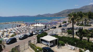 Ein Parkplatz an der Küste mit Autos, einer überdachten Parkgarage und einem Strand mit Sonnenschirmen und Personen im Hintergrund.