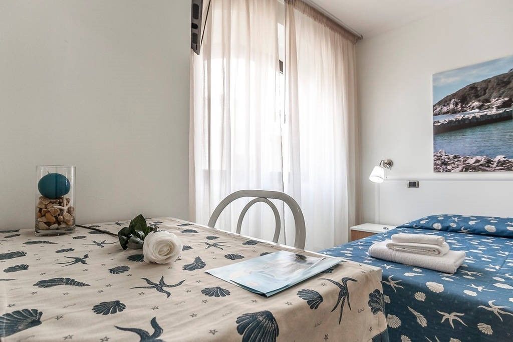 A bedroom with a bed, a table with a vase and a book, a chair, and a window with curtains.