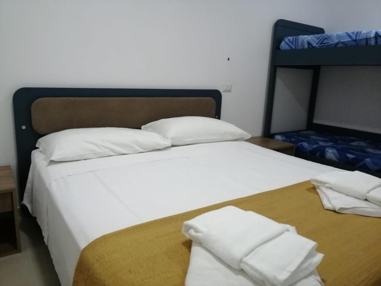 A single bed with white bedding and a mustard-colored throw blanket, a nightstand, and a bunk bed with a blue patterned bedspread.
