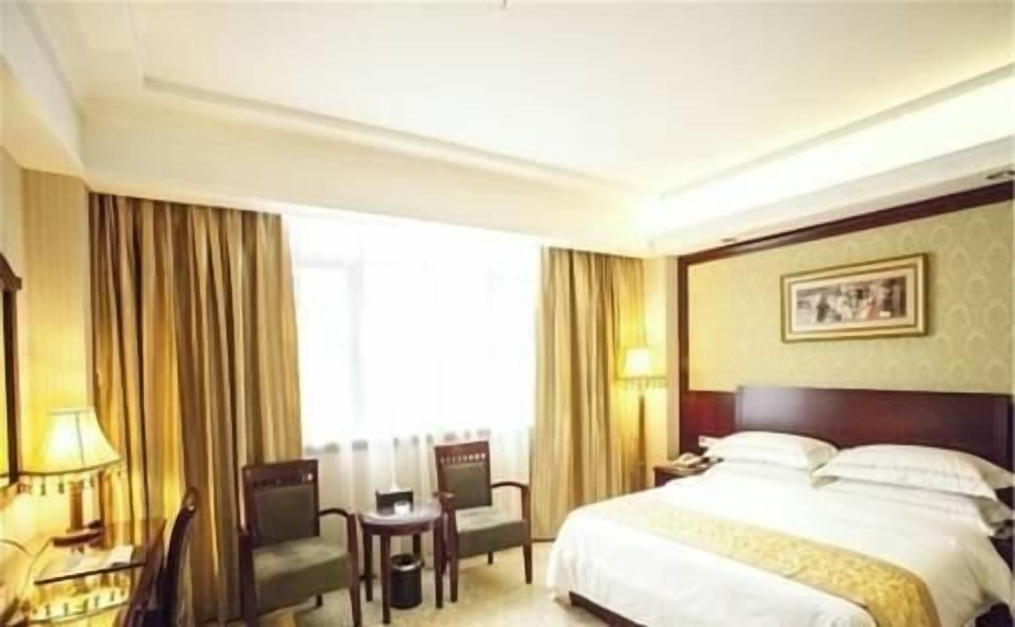 A hotel room with a bed, two chairs, a small table, a lamp, and a window with curtains.