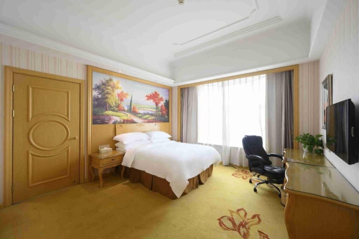 A hotel room with a bed, a desk, a chair, a TV, a painting, and a door.