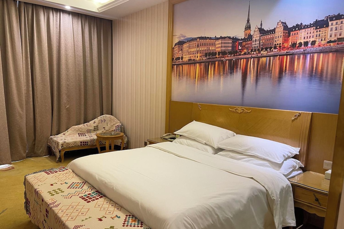 A hotel room with a large bed, a chair, a bedside table, and a painting of a cityscape.