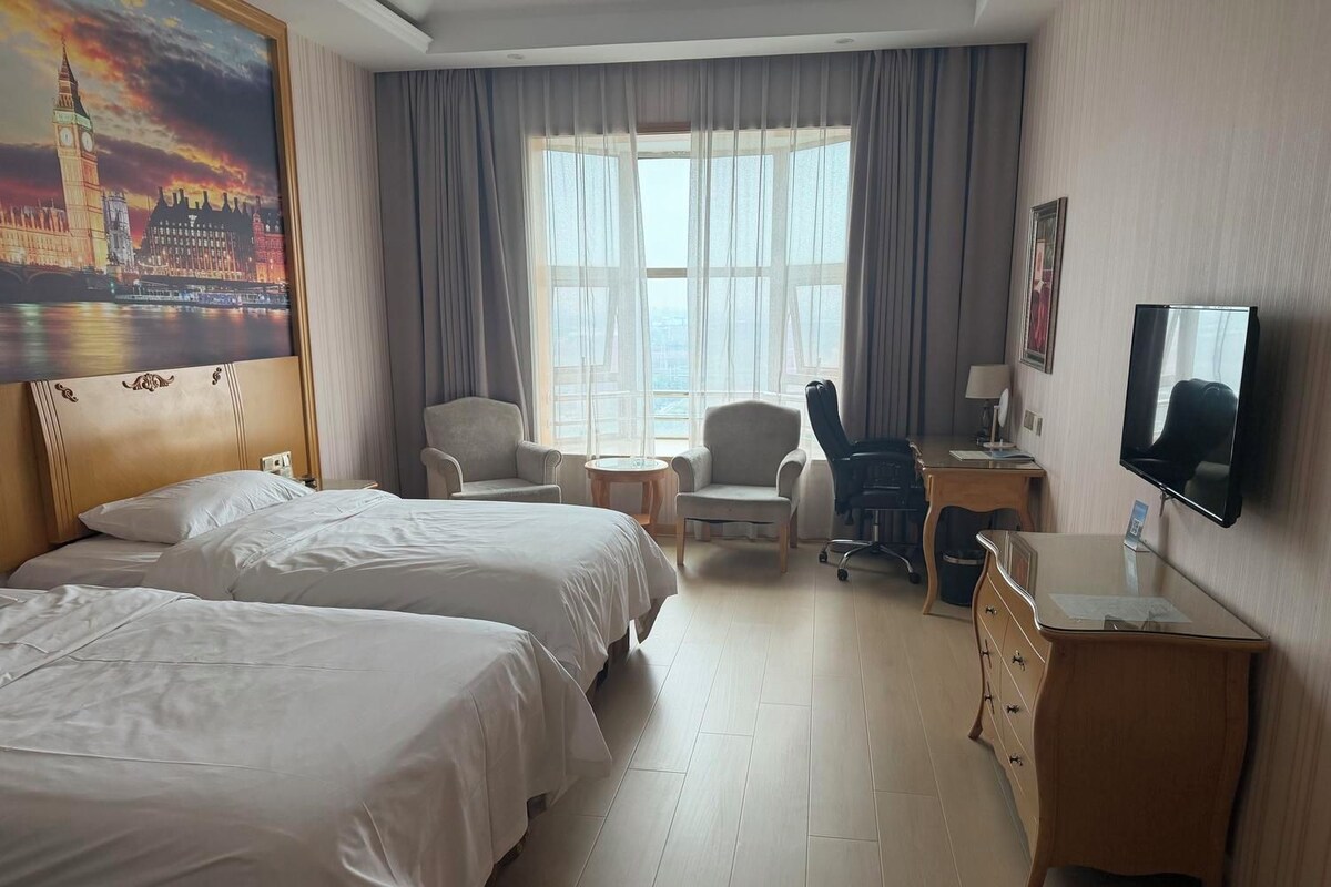 A hotel room with two beds, a desk, a chair, a television, and a large window.