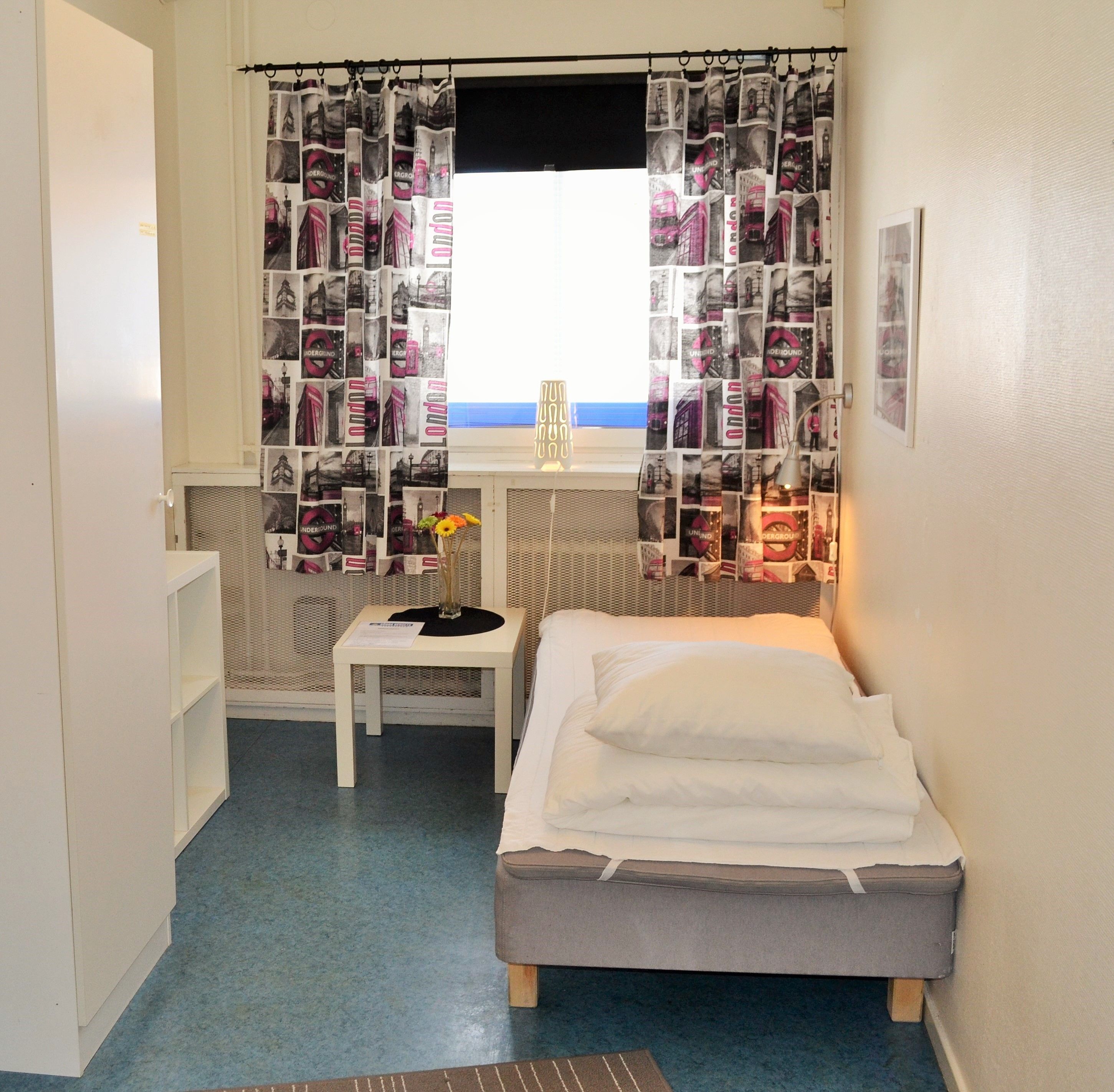 A small, compact room with a bed, a table, and a chair. A window with a curtain featuring a cityscape design is visible.