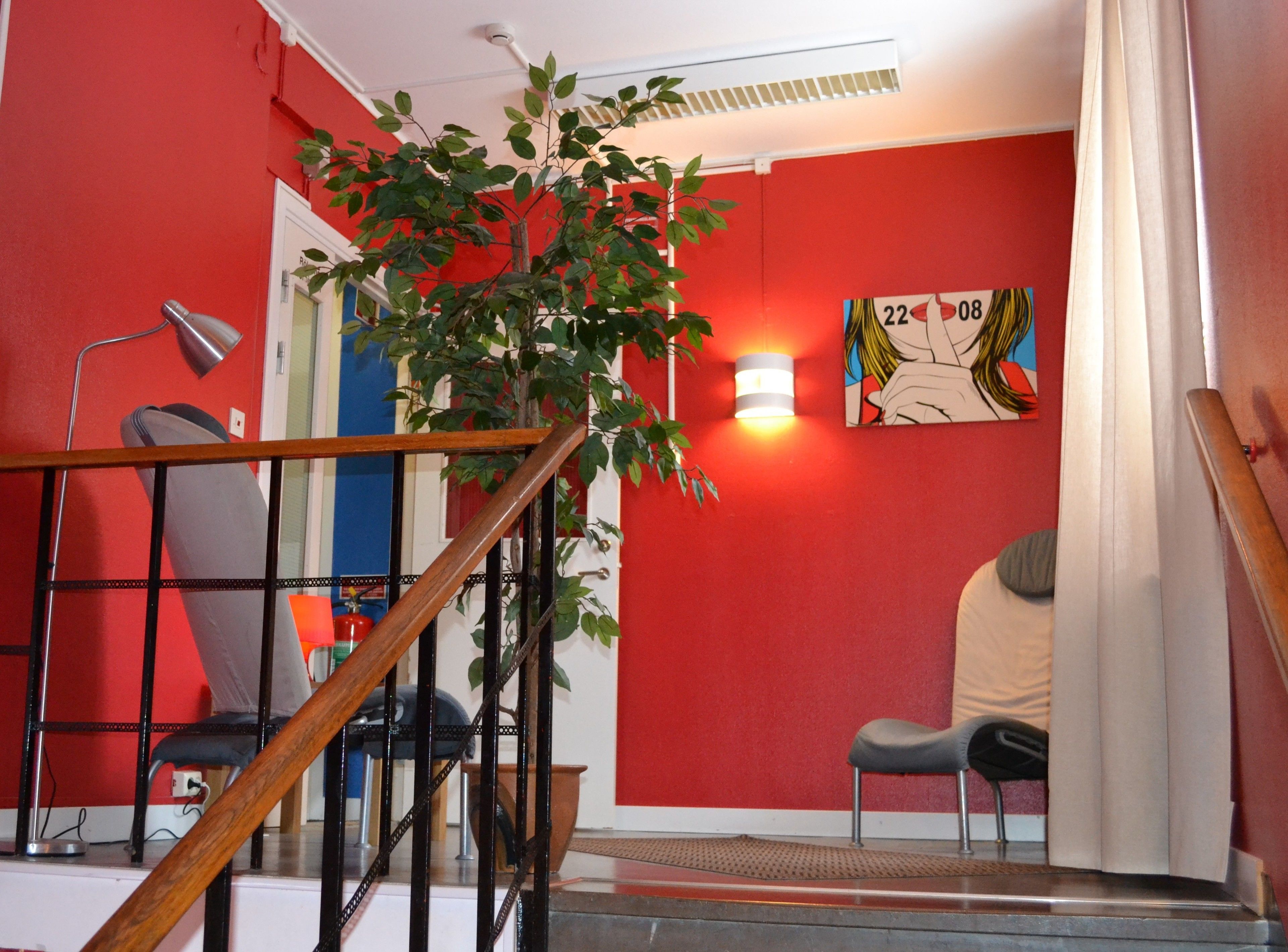 Un escalier avec une rampe en bois mène à une pièce aux murs rouges, où se trouvent une plante en pot, un tableau et une chaise.