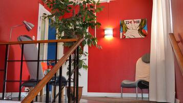 Un escalier avec une rampe en bois mène à une pièce aux murs rouges, où se trouvent une plante en pot, un tableau et une chaise.