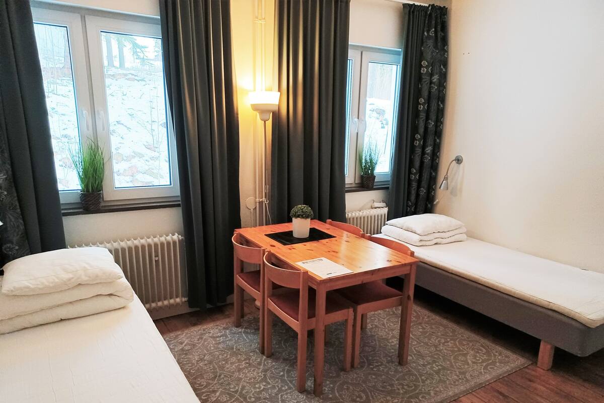 Une petite pièce accueillante, avec un lit, un canapé, une table et une lampe. On y trouve des fenêtres avec des rideaux, un radiateur et une plante sur le rebord de la fenêtre.