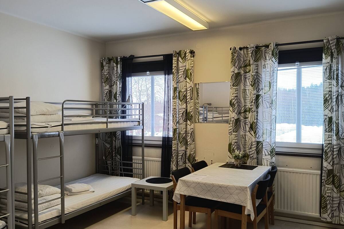 Une chambre de dortoir avec des lits superposés, une table à manger, des chaises, un tapis à motifs et de grandes fenêtres avec des rideaux.