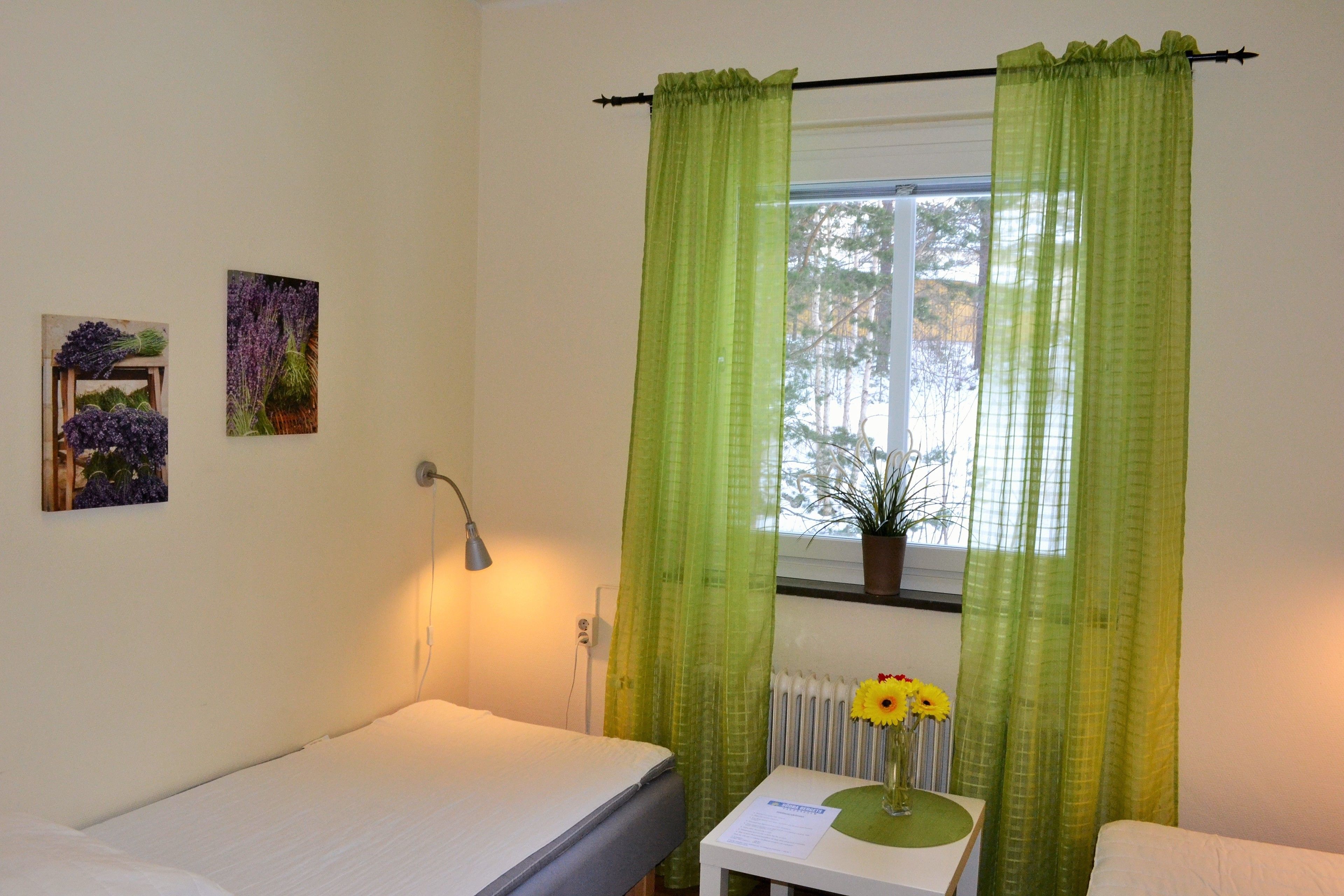 A room with a bed, a small table with a vase of flowers, and a window with green curtains.