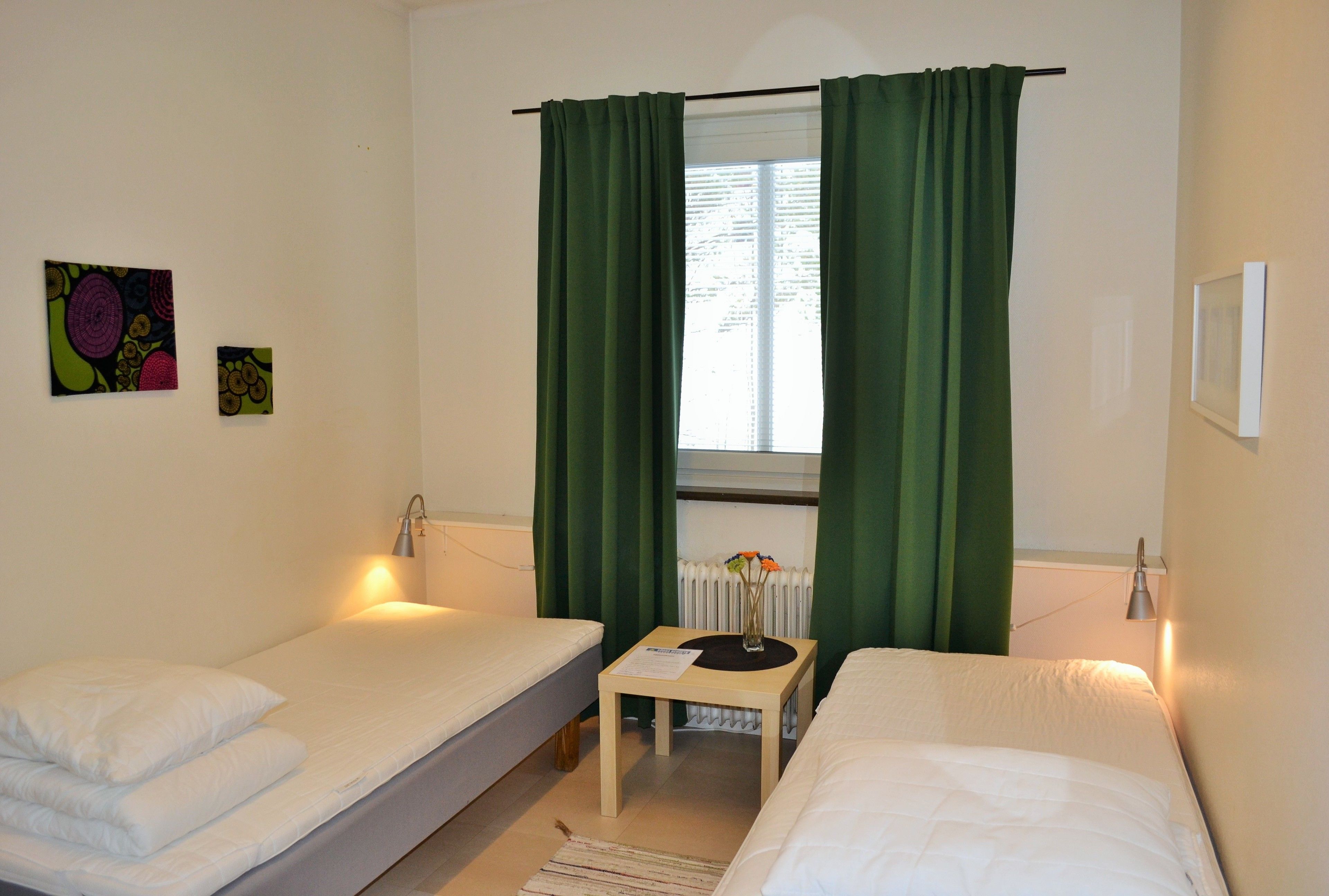 Una habitación pequeña y limpia con dos camas individuales, una mesita con un florero y una ventana con cortinas verdes.