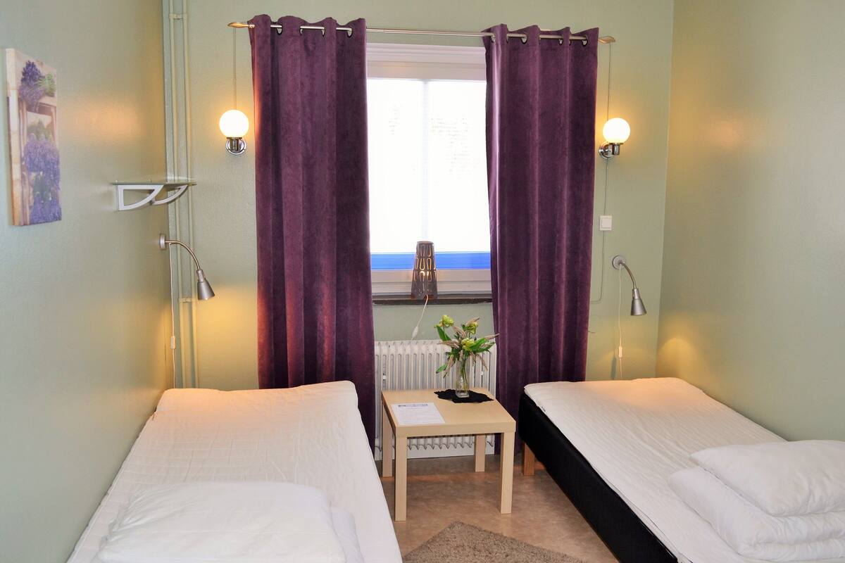 Une chambre avec deux lits simples, une petite table sur laquelle est posé un vase, et une fenêtre ornée de rideaux violets.
