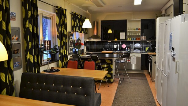 Una cocina compacta con zona de comedor, que presenta un cortinero con estampado de hojas negras y amarillas, una mesa de comedor de madera y un sofá negro.