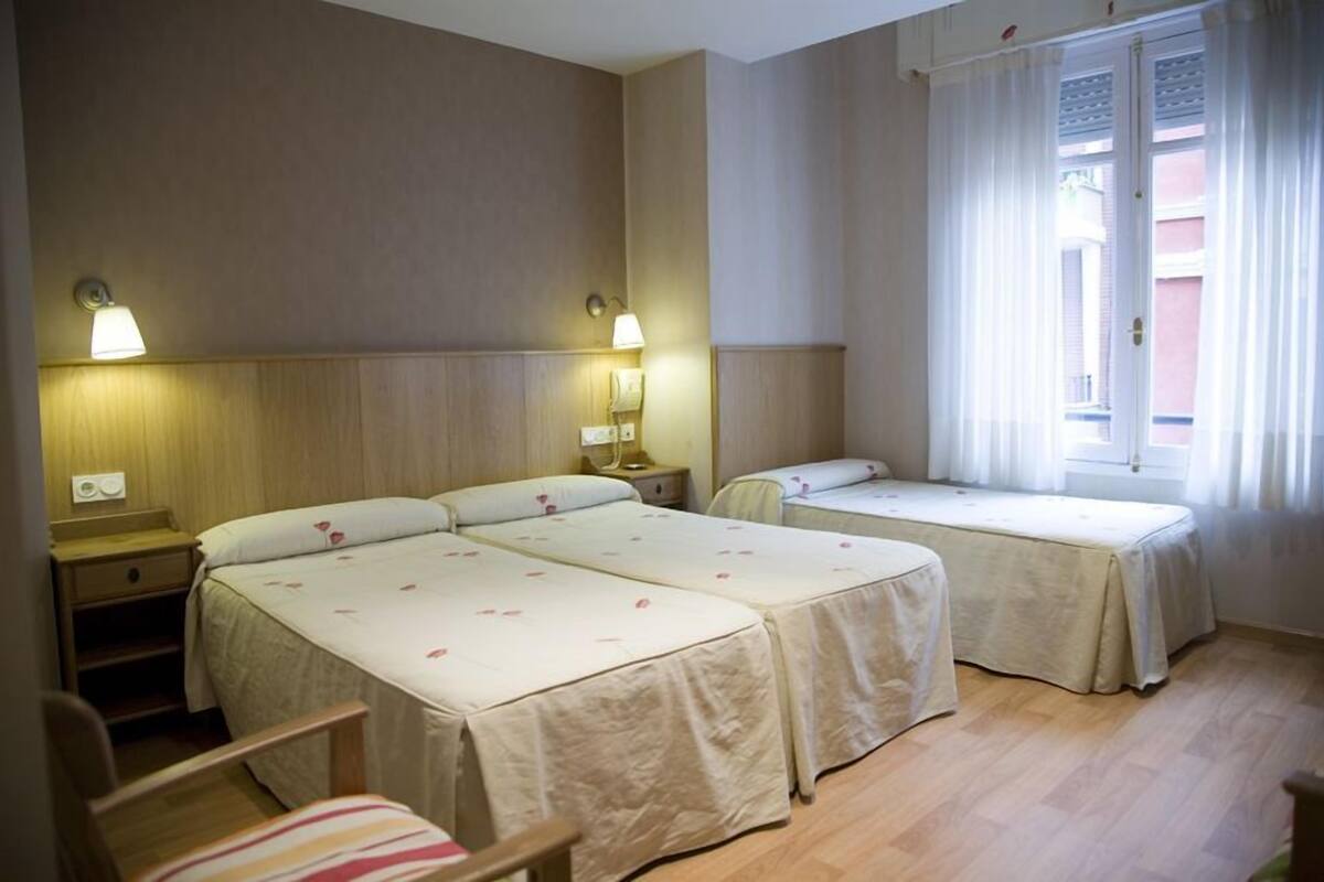 Quarto de hotel com duas camas de solteiro, luminárias de cabeceira, uma cadeira e uma janela com cortinas.