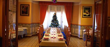 Un coin repas avec une longue table dressée pour le repas, entourée de chaises, d’un sapin de Noël et d’une fenêtre avec des rideaux.
