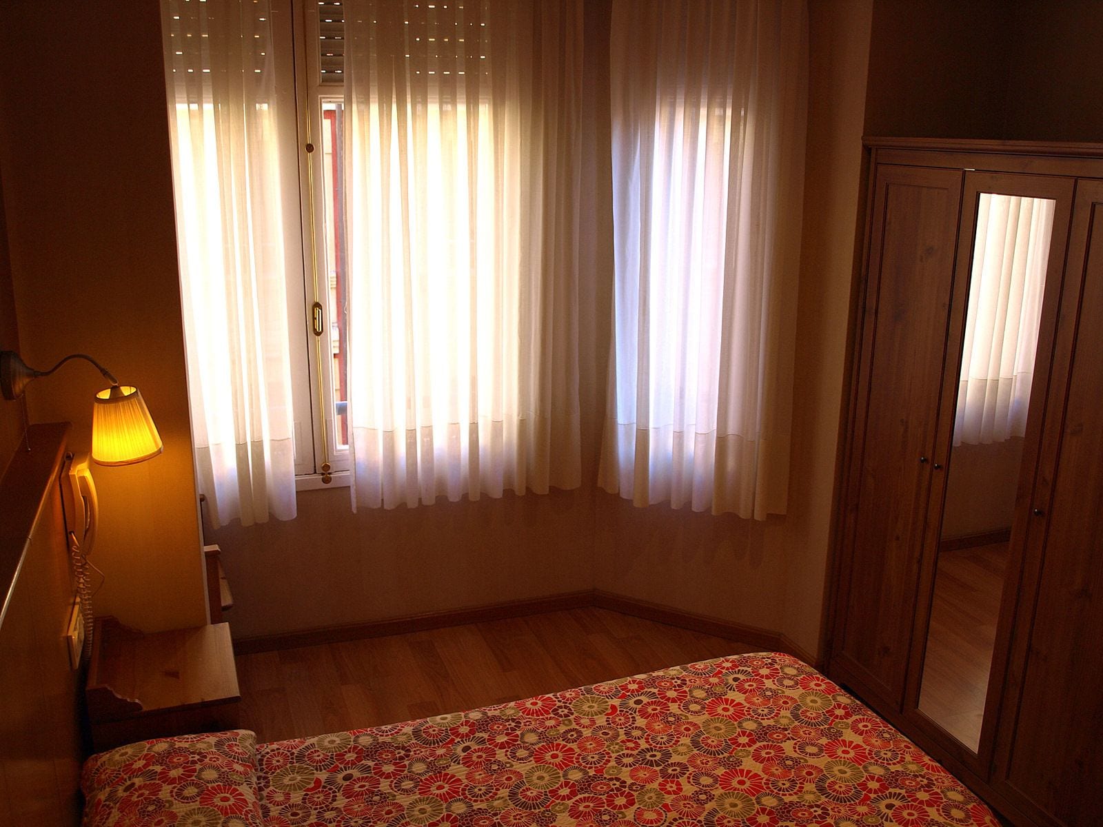 A bedroom with a bed, a lamp, a wardrobe, and a window with sheer curtains.