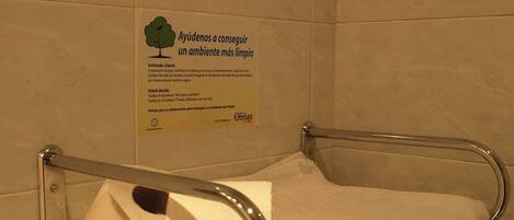 A bathroom with a handrail and a towel rack. A sign on the wall reads "Ayúdenos a conseguir un ambiente más limpio" in Spanish.
