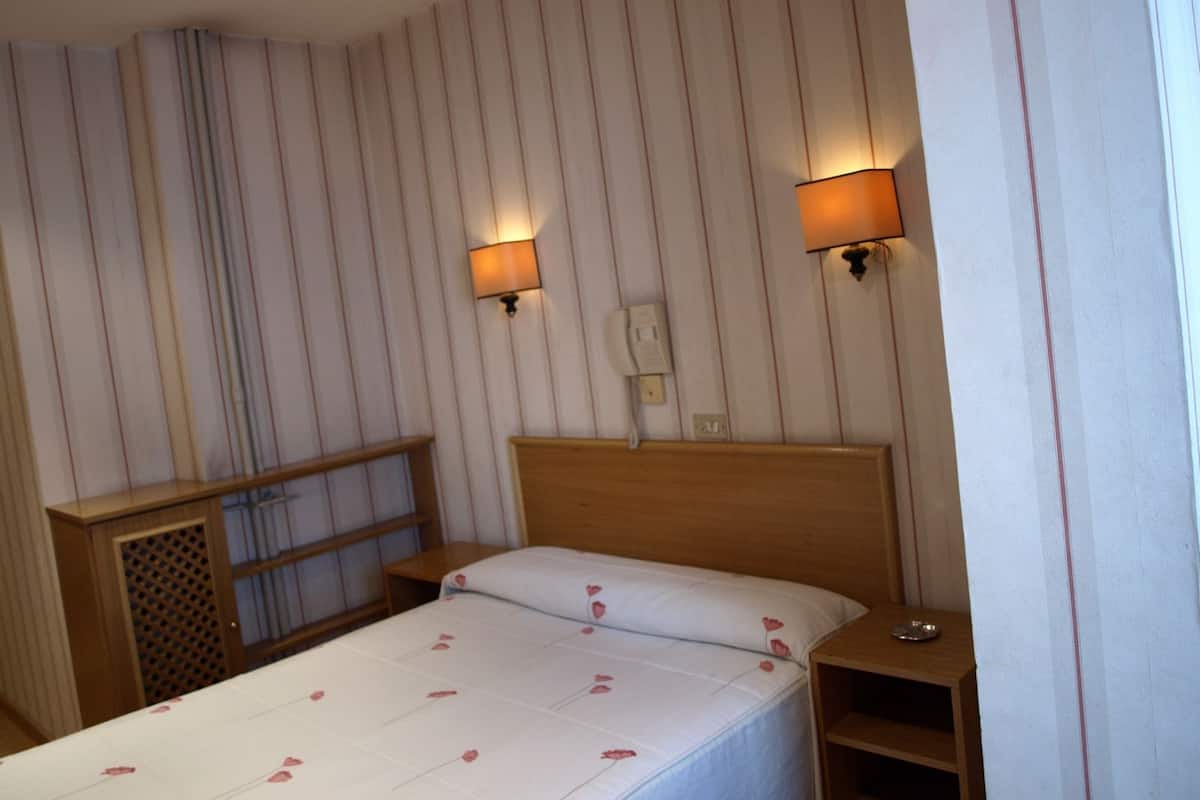 Quarto de hotel com uma cama, duas mesas de cabeceira, um painel de cama de madeira, um telefone e duas luminárias fixadas na parede.