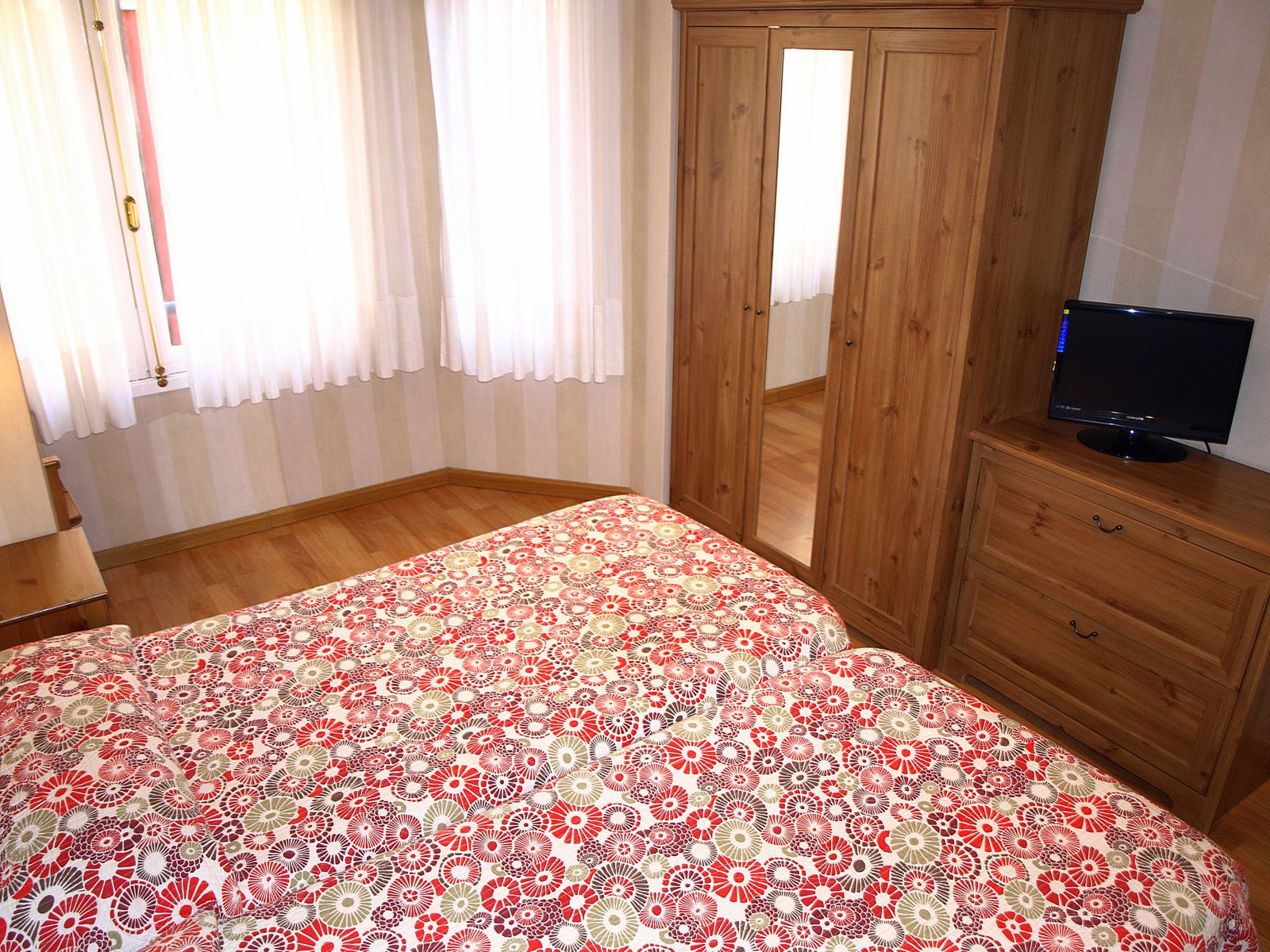 Une chambre à coucher avec un lit, une armoire en bois, un téléviseur posé sur une commode et une fenêtre avec des rideaux.