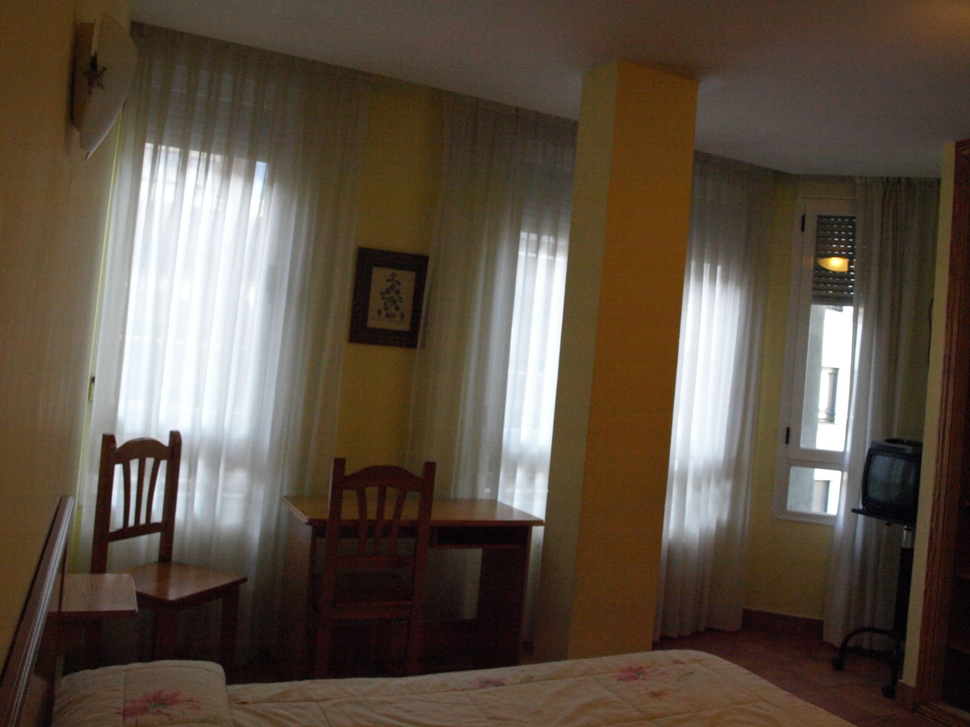 Habitación de hotel con cama, una silla de madera, una mesita pequeña y un escritorio. Se ve una ventana con cortinas, un cuadro en la pared y un televisor sobre un mueble.