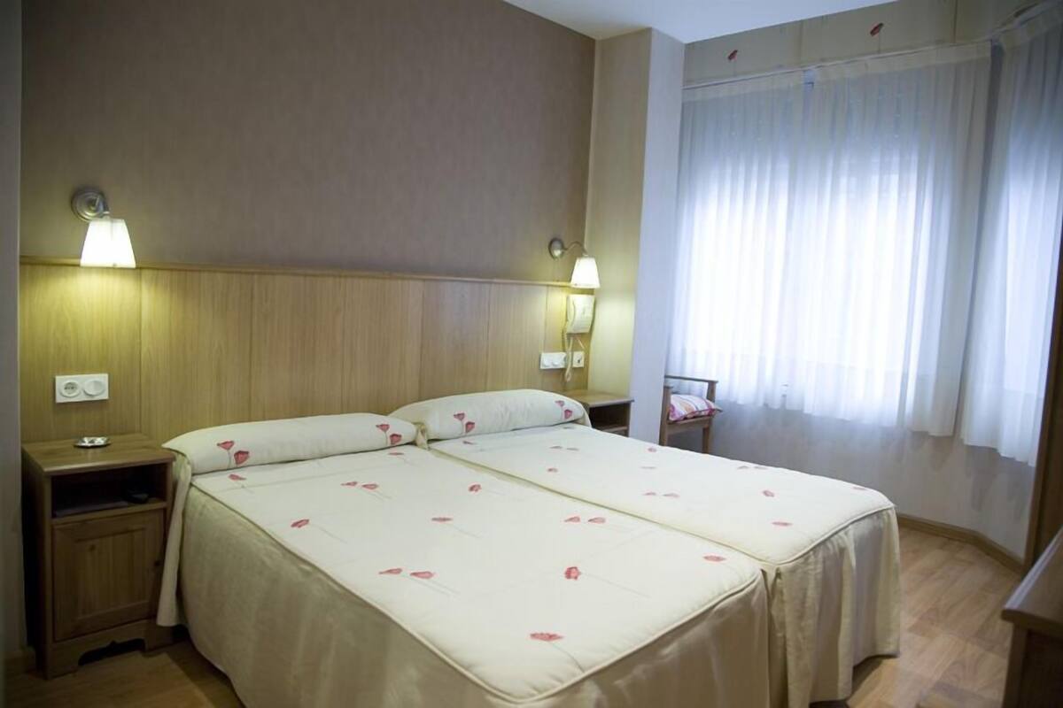 Quarto de hotel com cama de casal, duas mesas de cabeceira, um criado-mudo, uma lâmpada e uma janela com cortinas.